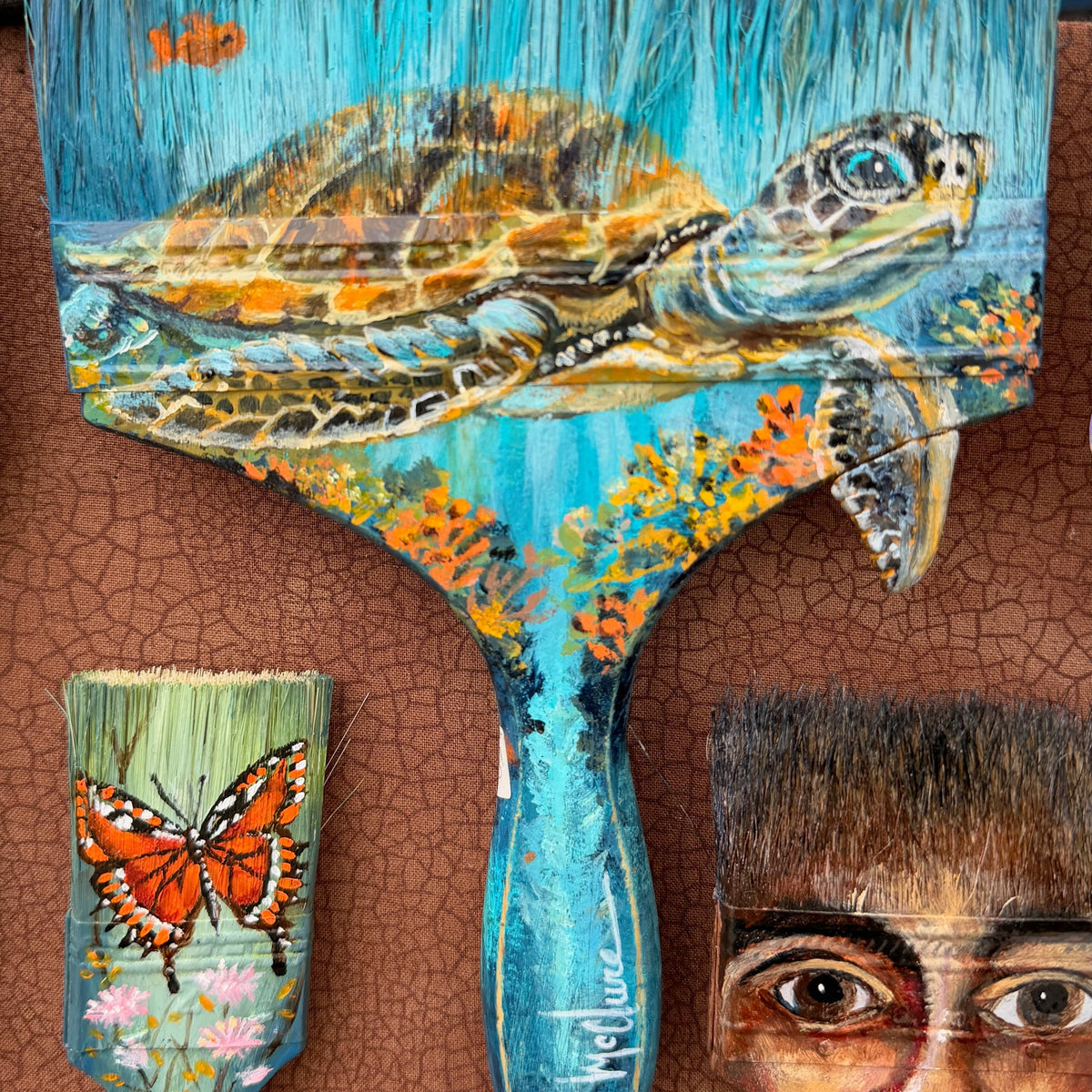 A decorative hand-painted wooden paintbrush with a sea turtle design, displayed alongside other painted brushes on a brown textured wall.
