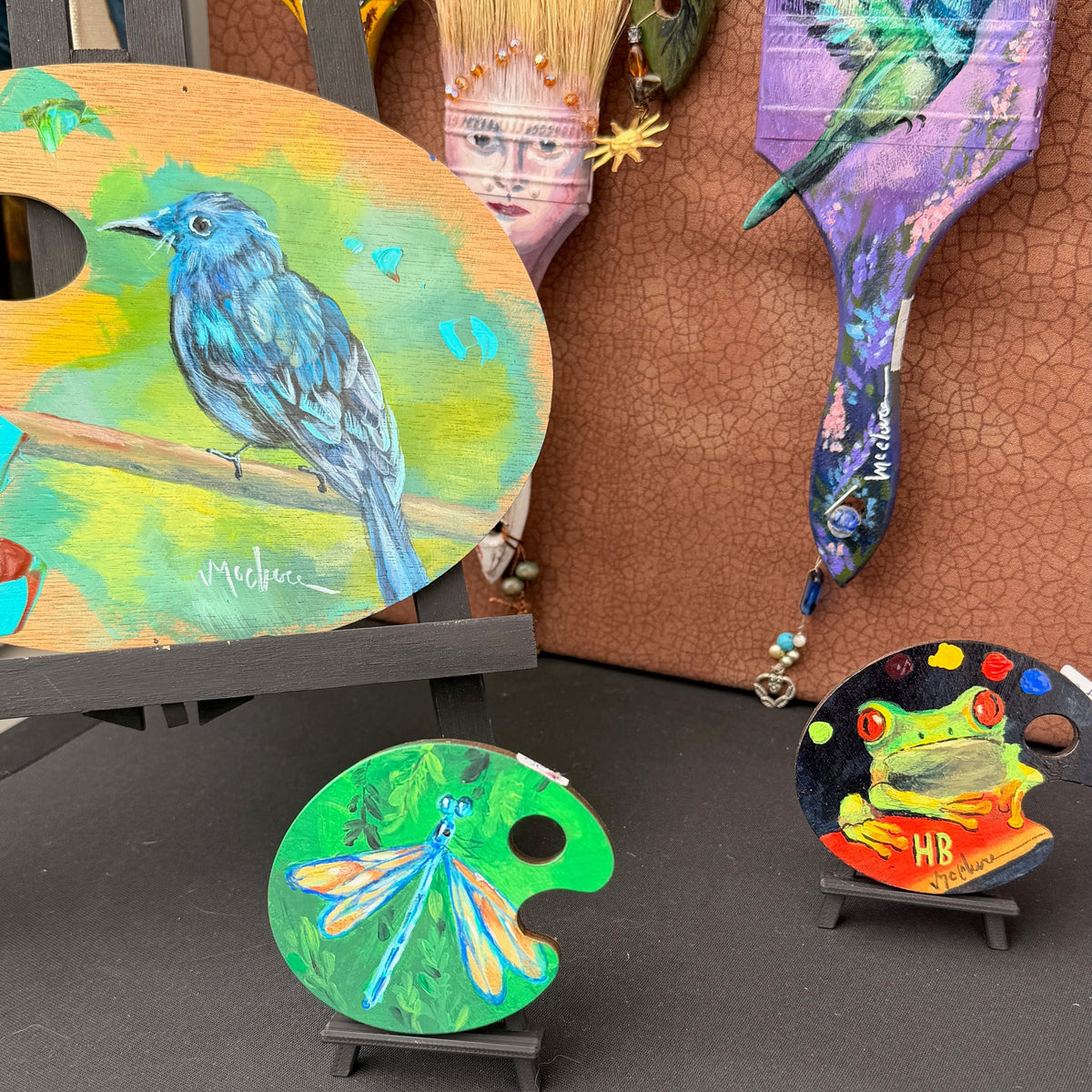 A collection of colorful hand-painted wooden art pieces displayed on stands, including a large bird-themed painter's palette, a dragonfly-themed palette, a frog-themed palette, and several decorative paintbrushes with painted bristles against a brown textured wall.